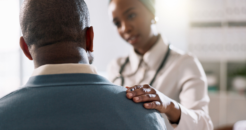 Healthcare professional places a hand on a patient's shoulder in a comforting exam room setting.