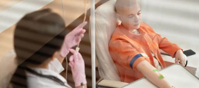Patient in an orange hospital gown sits in bed with an IV in their arm; a clinician in a mask stands outside behind glass, preparing a procedure.