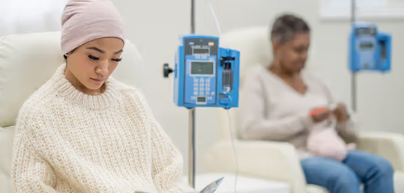 Woman with a headscarf sits in a sweater beside an IV pole in a hospital room; another patient reads in the background.