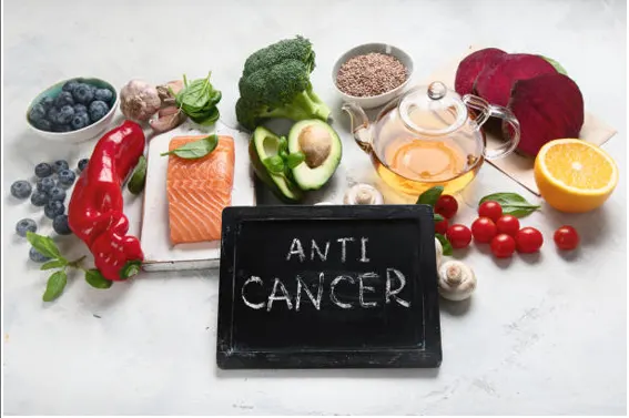 Assortment of healthy foods (blueberries, red pepper, salmon, broccoli, avocado, beets, citrus) around a chalkboard reading ANTI CANCER.