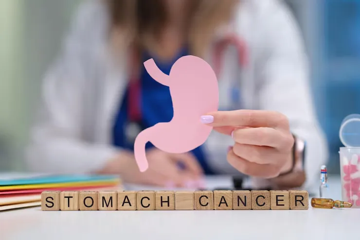 Medical professional holds a pink stomach cutout over a desk with wooden blocks spelling 'STOMACH CANCER'.